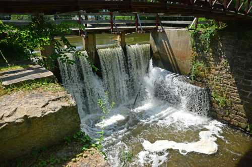 A small waterfall cascades over a dam, surrounded by greenery and rocky terrain under a clear sky.