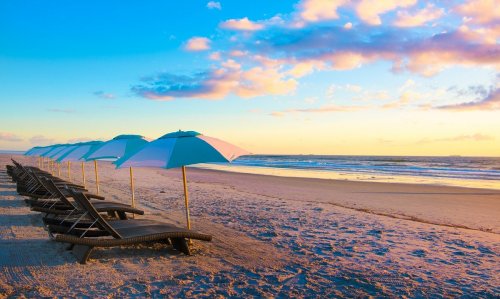 Beach scene at sunset with lounge chairs and blue umbrellas lining the shore, reflecting colorful skies.
