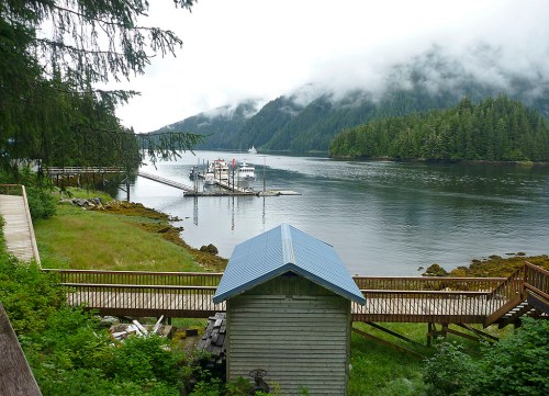 A serene waterfront scene with a dock, surrounded by lush greenery and misty mountains in the background.