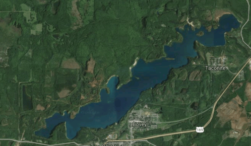 Aerial view of a large lake surrounded by dense forest and small towns, with roads visible nearby.