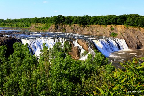 A scenic view of a waterfall cascading over rocky cliffs, surrounded by lush green trees and a river below.