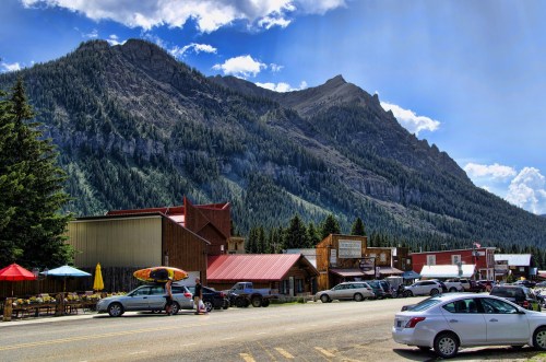 A scenic mountain town with shops, parked cars, and outdoor seating under a blue sky with clouds.