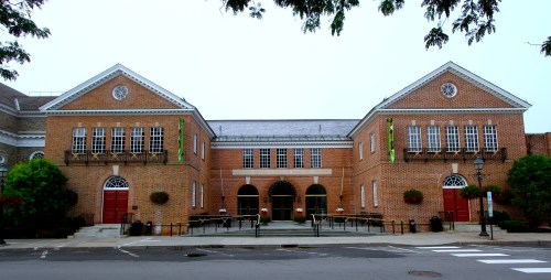 A brick building with arched windows and red doors, surrounded by greenery and a sidewalk.