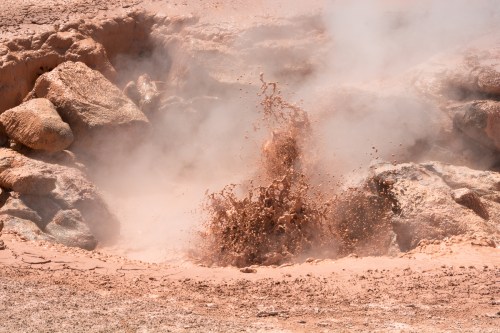 A geyser erupts, sending mud and steam into the air amidst rocky terrain.