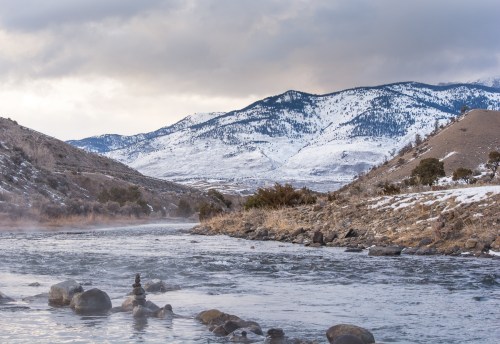 A serene river flows through a snowy landscape, with mountains in the background under a cloudy sky.