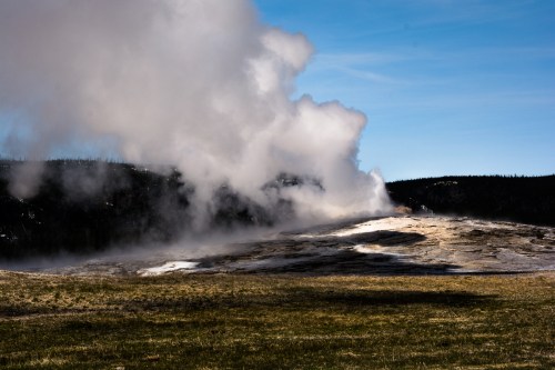 A geyser erupts steam into the air against a backdrop of green hills and a clear blue sky.