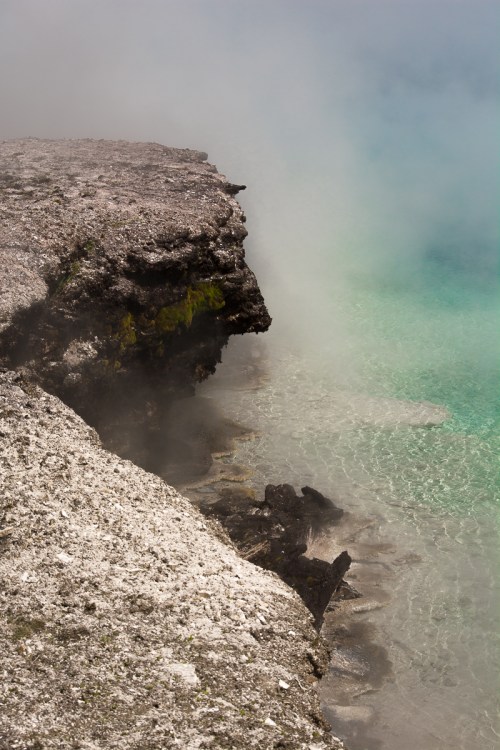 A rocky shoreline with steam rising from the water, revealing clear turquoise water and a misty atmosphere.