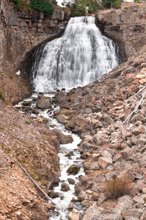 A cascading waterfall flows over rocky terrain, surrounded by rugged cliffs and sparse greenery.