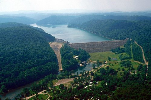 Aerial view of a dam surrounded by lush green hills and a serene lake, with a river flowing below.