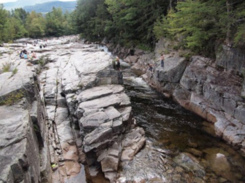A rocky riverbed with people exploring along the water's edge, surrounded by trees and mountains in the background.