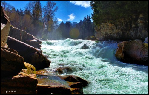 A rushing river flows over rocks, surrounded by trees and mountains under a bright blue sky.