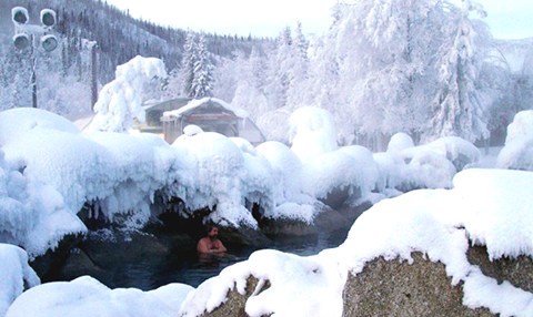 A person relaxes in a steaming hot spring surrounded by snow-covered trees and rocks in a winter landscape.