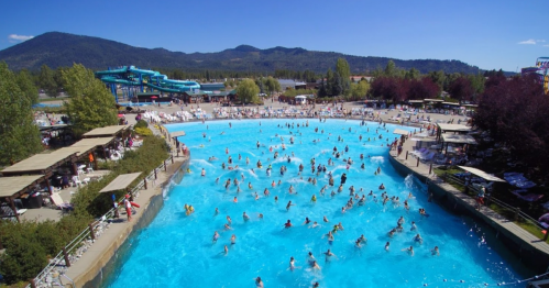 A crowded water park pool on a sunny day, with slides in the background and mountains in the distance.