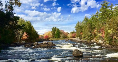 A scenic river flows through a colorful autumn landscape with trees in vibrant shades of red, orange, and green.