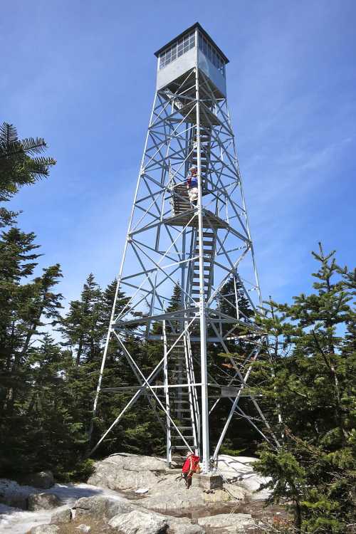 A tall metal fire tower surrounded by trees, with a person climbing the stairs and another at the base.