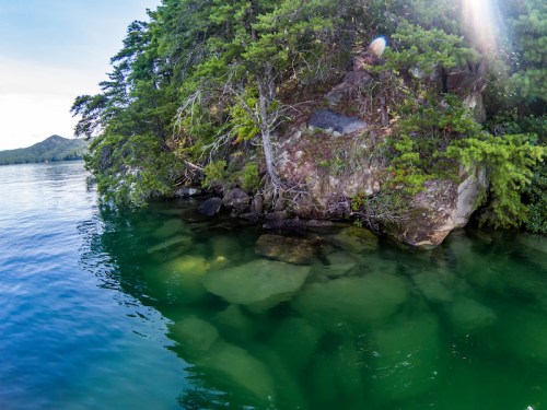 A serene lake scene with clear green water revealing rocks beneath, surrounded by lush trees and a rocky shoreline.