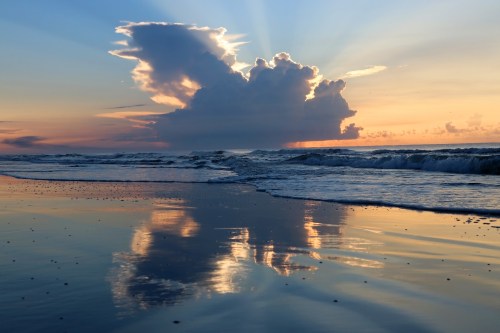 A serene beach scene at sunset, featuring clouds reflecting on calm water with gentle waves lapping at the shore.