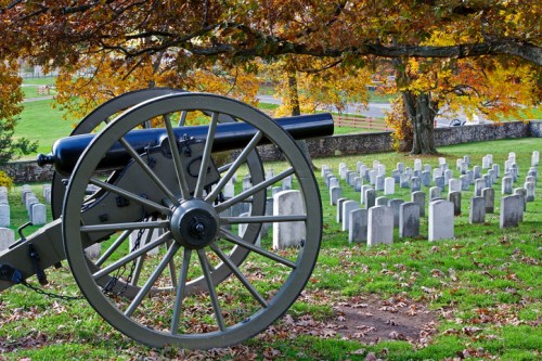 A historic cannon overlooks a cemetery with white gravestones, surrounded by vibrant autumn foliage.