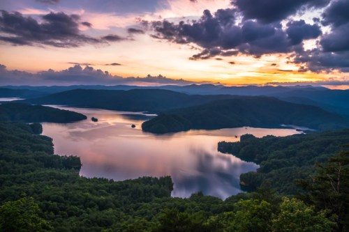A serene sunset over a lake surrounded by mountains and lush greenery, with colorful clouds reflecting on the water.