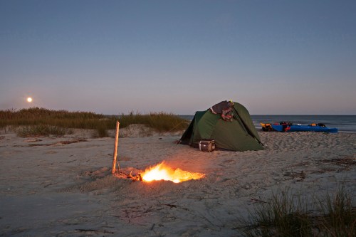 A tent by the beach at dusk, with a campfire glowing and kayaks in the background under a rising moon.