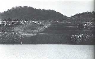 A black and white landscape showing a barren hillside with a body of water in the foreground and trees in the background.