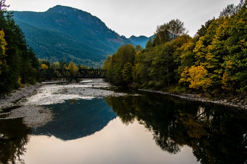 A tranquil river reflects mountains and autumn trees, with a bridge crossing in the background.