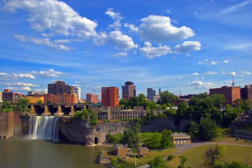 A scenic view of a waterfall with a city skyline and blue sky filled with clouds in the background.