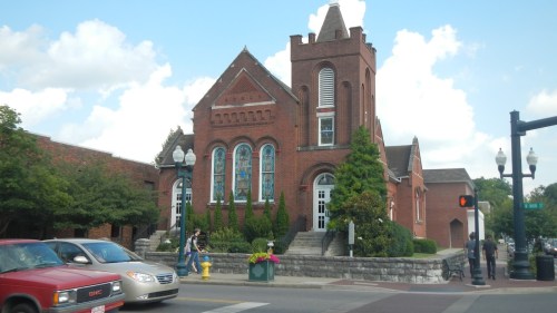 Historic brick building with tall windows, surrounded by greenery, on a busy street with cars and pedestrians.