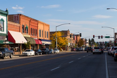 A bustling street lined with brick buildings, shops, and parked cars under a clear blue sky.