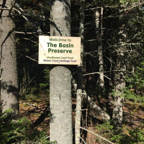 Wooden sign reading "Welcome to The Basin Preserve" among trees, indicating a protected area in Maine.