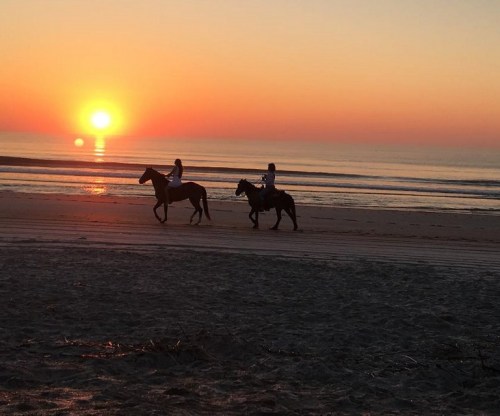 Two riders on horseback walk along a beach at sunset, with the sun setting over the calm ocean.