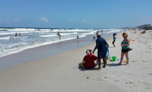 A sunny beach scene with people playing in the sand and water, enjoying a warm day by the ocean.