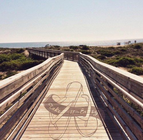 A wooden boardwalk leads to the beach, with the ocean visible in the background and a sunny sky above.