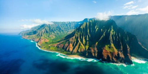 Aerial view of lush green mountains meeting the turquoise ocean, with waves gently crashing on the shore.