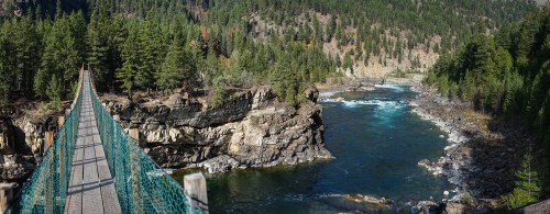 A suspension bridge spans a river surrounded by lush green trees and rocky cliffs under a clear blue sky.