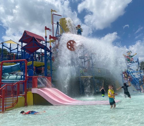 A water park scene with kids playing in a splash area, featuring slides and a large water feature. Bright, sunny day.