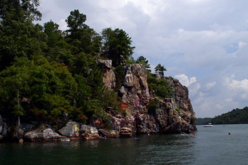 A rocky shoreline with trees, colorful graffiti, and a calm lake under a cloudy sky.
