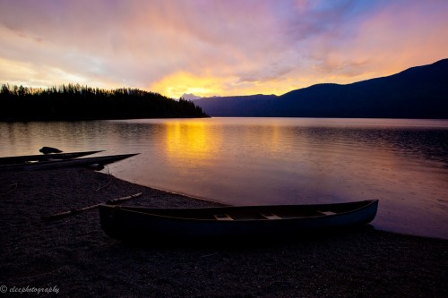 A serene lake at sunset, with a canoe on the shore and mountains silhouetted against a colorful sky.