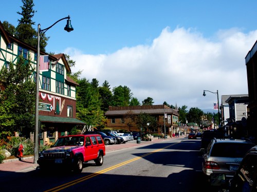 A quaint street lined with shops and trees, featuring a red vehicle and a blue sky with clouds.