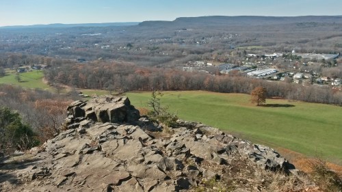 A rocky overlook with a panoramic view of a valley, fields, and distant hills under a clear blue sky.