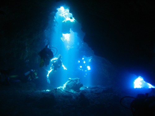 Divers explore an underwater cave illuminated by beams of sunlight filtering through the water.