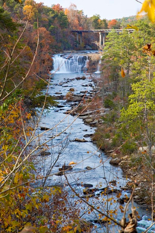 A scenic view of a waterfall surrounded by autumn foliage, with a bridge in the background over a flowing river.