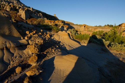 A rugged landscape with sandy hills and rock formations under a clear blue sky, casting long shadows in the evening light.