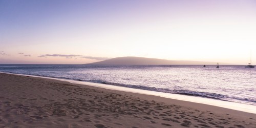 A serene beach at sunset, with gentle waves and distant mountains under a pastel sky.