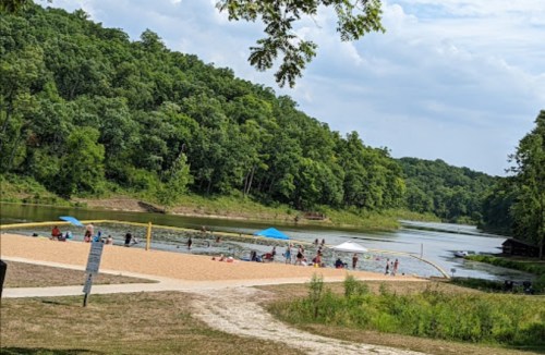A sandy beach area by a river, with people swimming and relaxing under umbrellas, surrounded by lush green trees.
