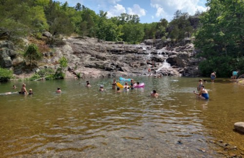 People swimming and playing in a natural pool near a waterfall, surrounded by lush greenery and rocky terrain.
