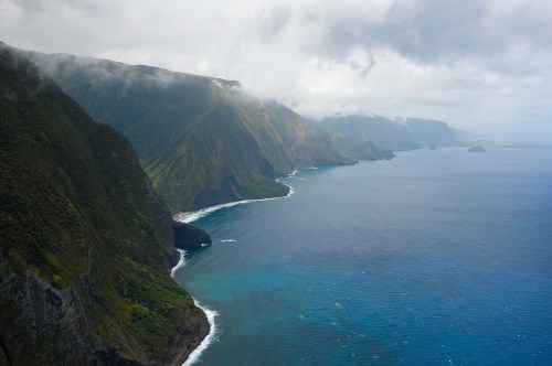 A dramatic coastline with steep cliffs and lush greenery, meeting a calm blue ocean under a cloudy sky.
