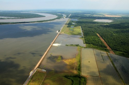 Aerial view of a flooded landscape with a road, river, and green fields under a cloudy sky.