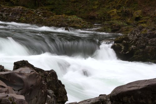 A flowing river cascades over rocks, creating a smooth, white water effect amidst a lush, green landscape.