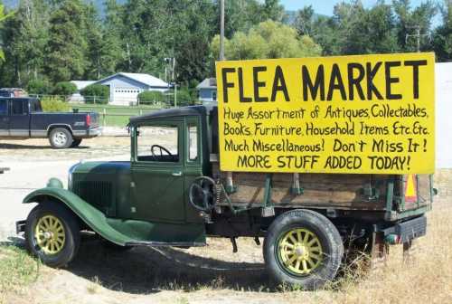 A vintage truck parked beside a large sign advertising a flea market with various items for sale.